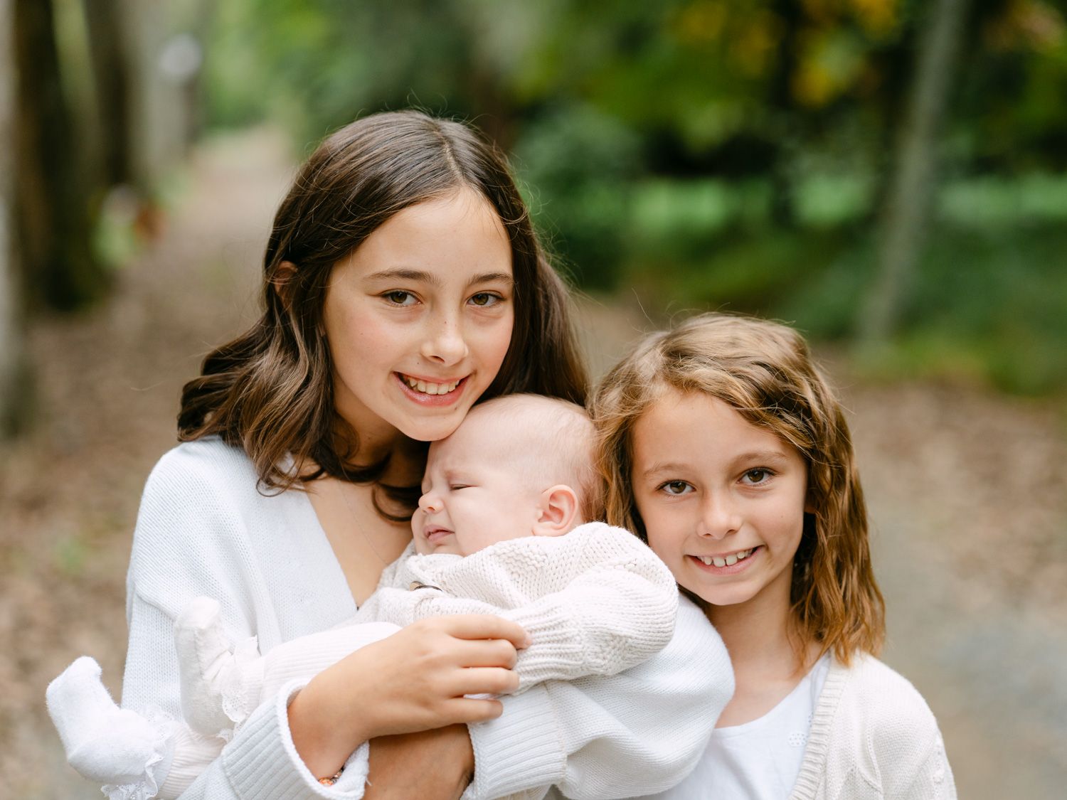 Two sisters in white sweaters lovingly hold their newborn sibling outdoors in a lush green forest setting.
