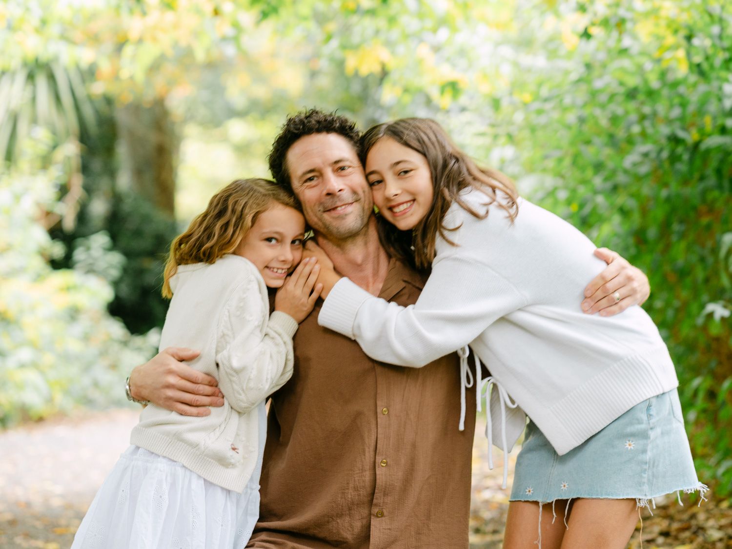 A man smiles warmly while being hugged by two young girls outdoors in a lush green garden setting.