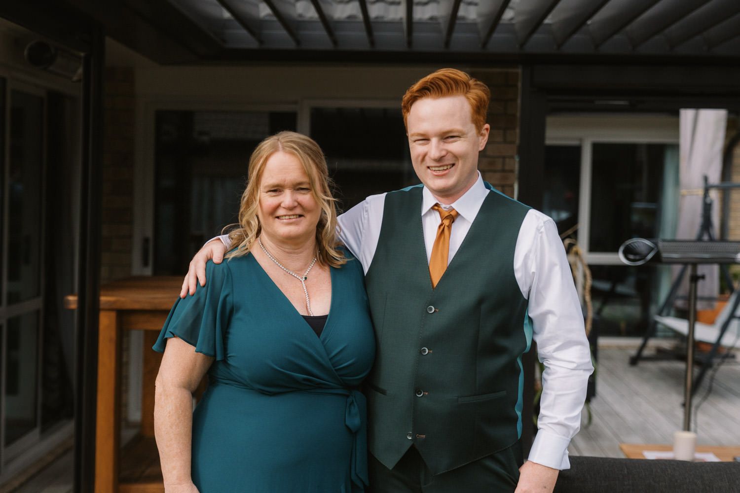 A smiling woman in a teal dress and a young redheaded man in a green vest and gold tie pose together outdoors.