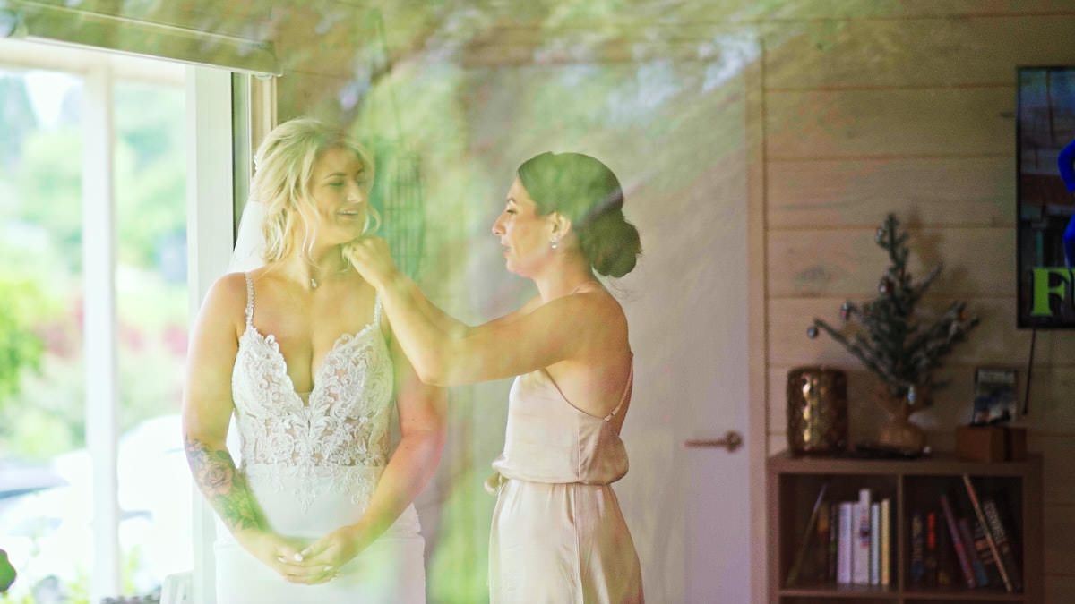 Bridesmaid adjusting bride's hair in a sunlit room, bride wearing elegant lace wedding dress.
