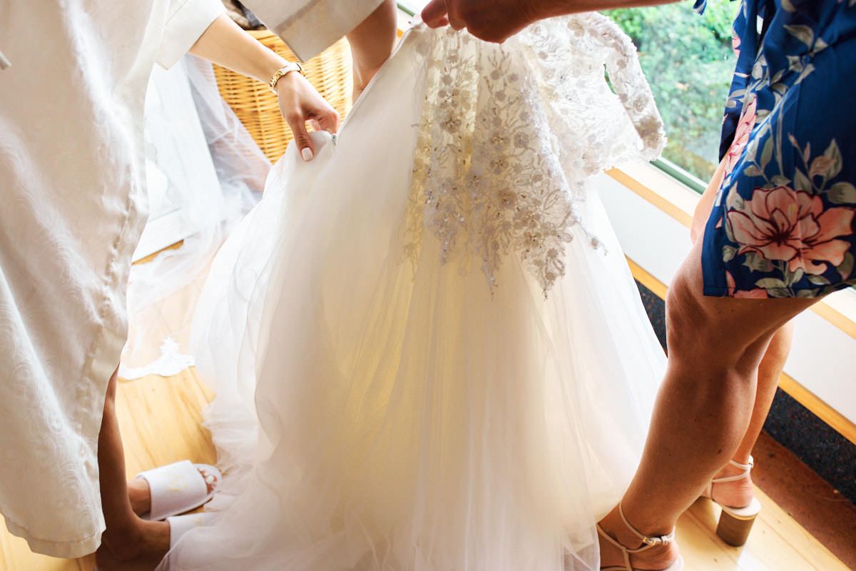 Bride being helped into an ornate beaded white wedding gown by two attendants near a window.