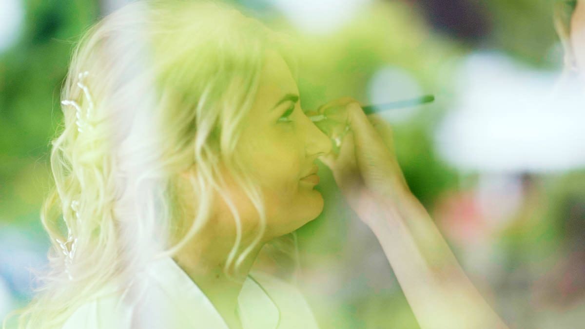 A makeup artist applying makeup to a blonde woman outdoors in soft green natural light.