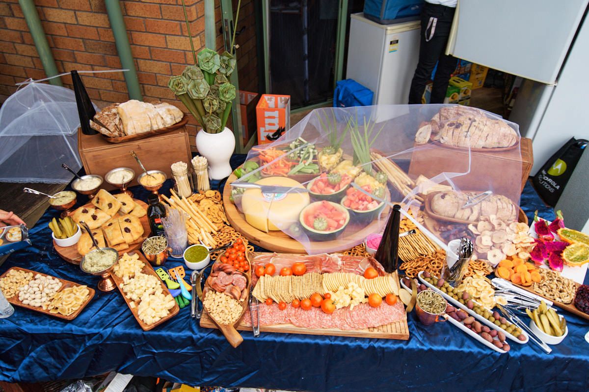 Elaborate charcuterie board spread on blue velvet table with meats, cheeses, crackers, and snacks at an outdoor party.