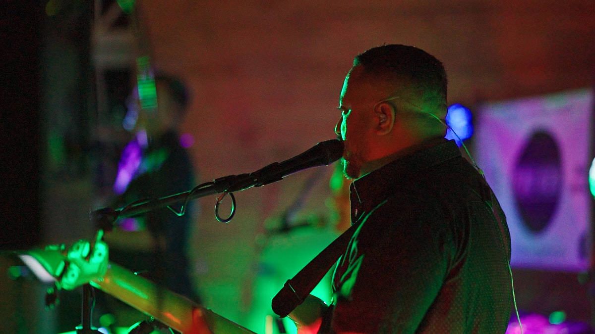 A male vocalist performs on stage with a microphone, bathed in green and pink stage lights during a live concert.