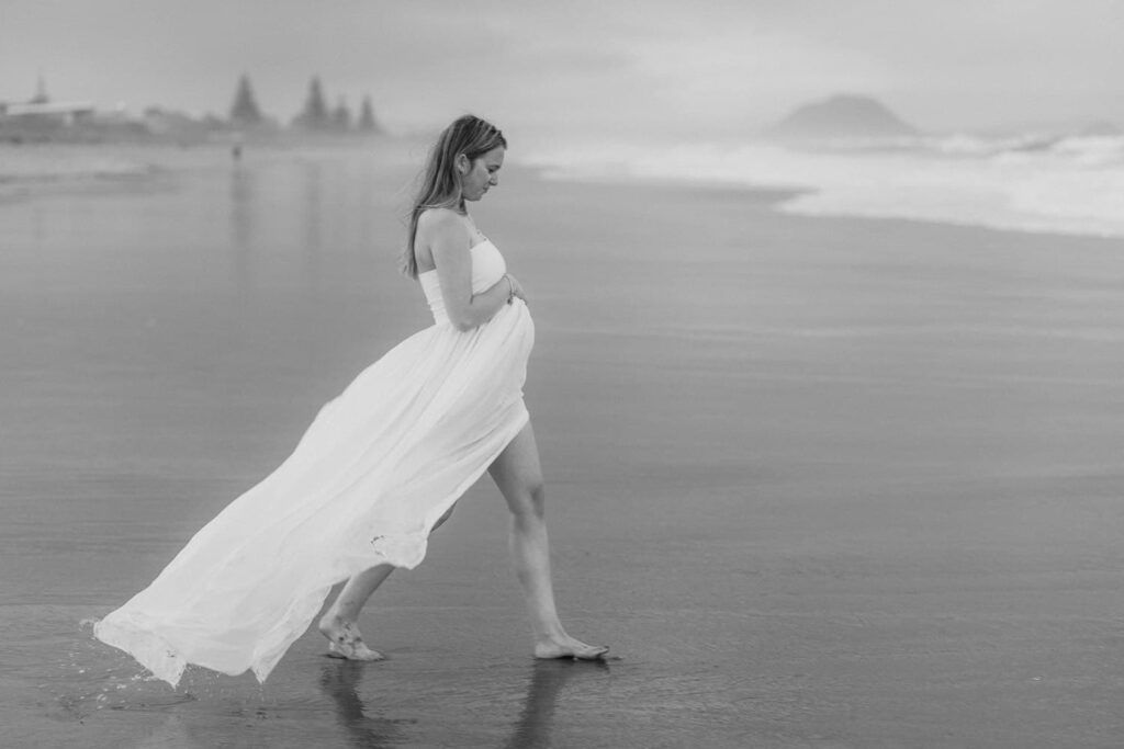 Pregnant woman in flowing white dress walking barefoot on beach in black and white maternity photo.