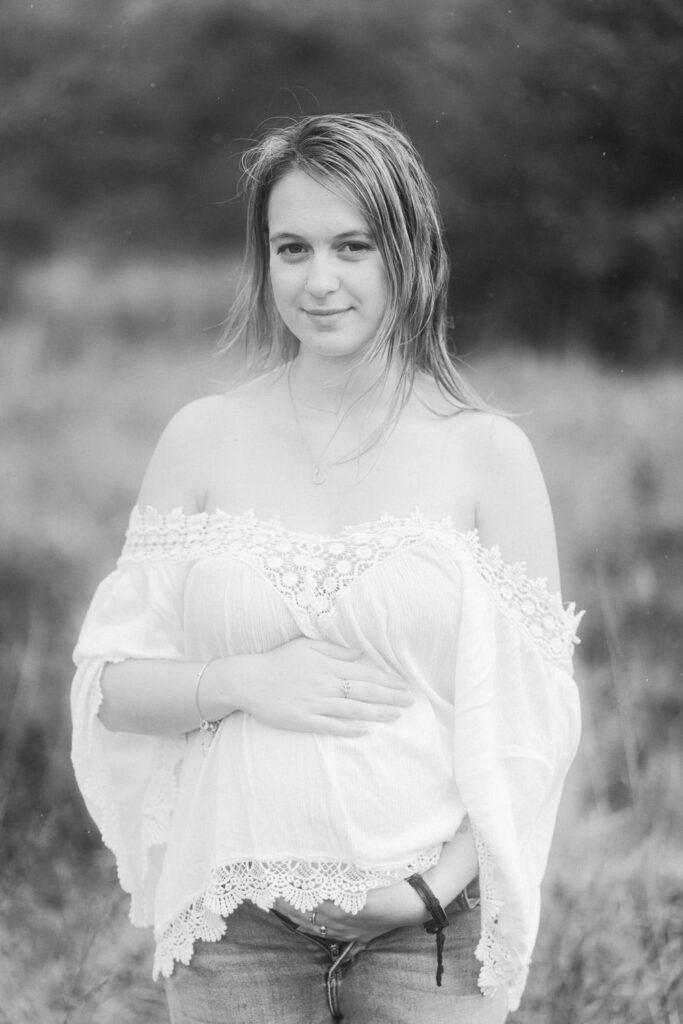 Black and white portrait of a young woman in an off-shoulder white lace top and jeans posing outdoors in a field.
