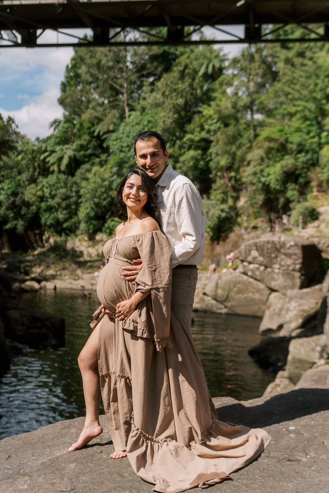 Pregnant couple posing outdoors near a river, woman in flowing beige dress, man in white shirt, lush greenery background.