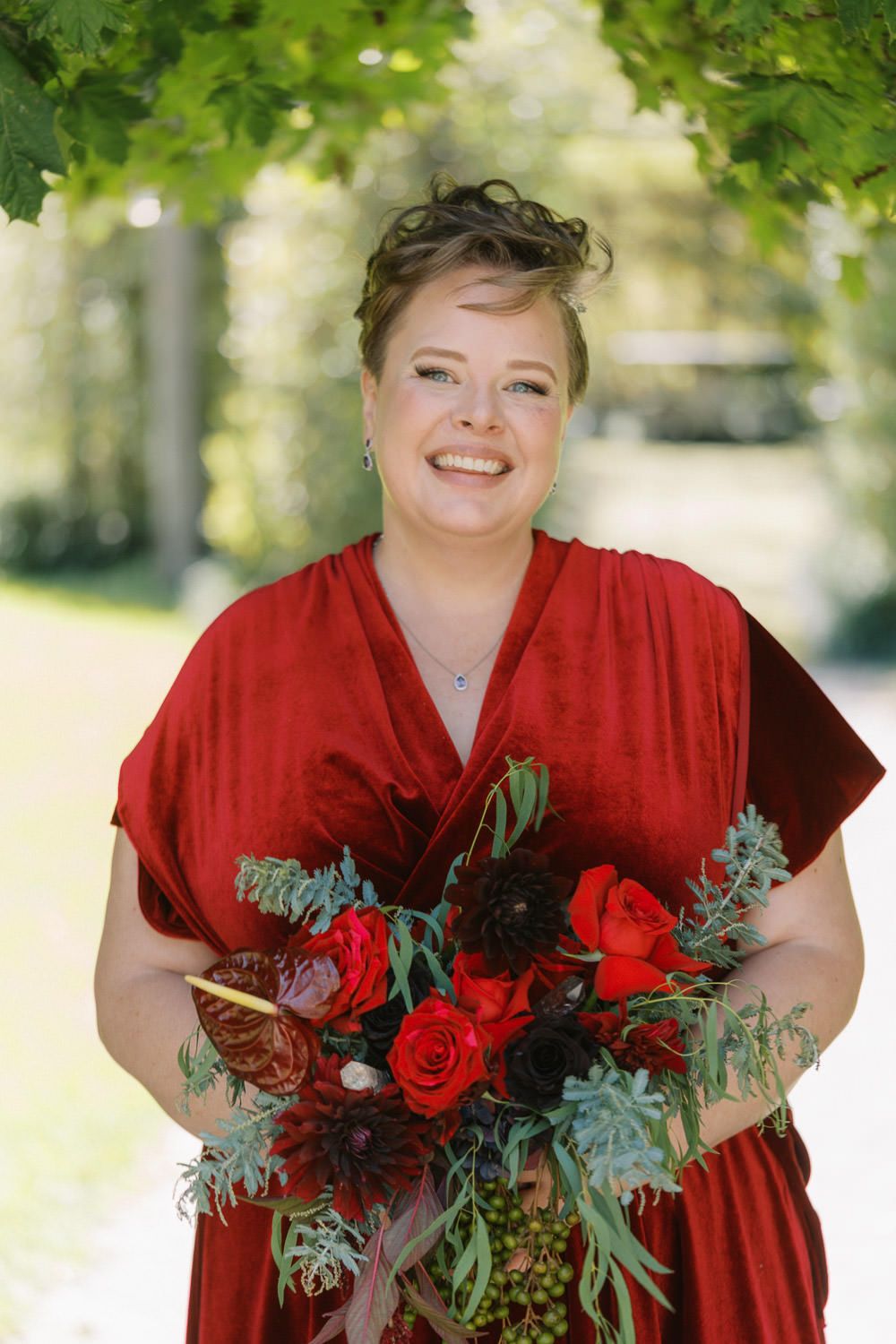 Smiling woman in red dress holding bouquet of red roses and greenery outdoors at a wedding celebration.