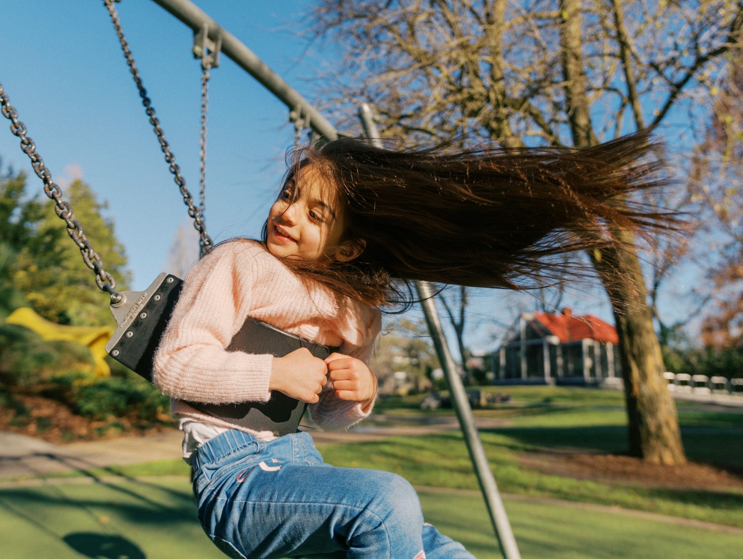 Young girl with flowing dark hair enjoys a sunny day on a playground swing, wearing a pink sweater and jeans.