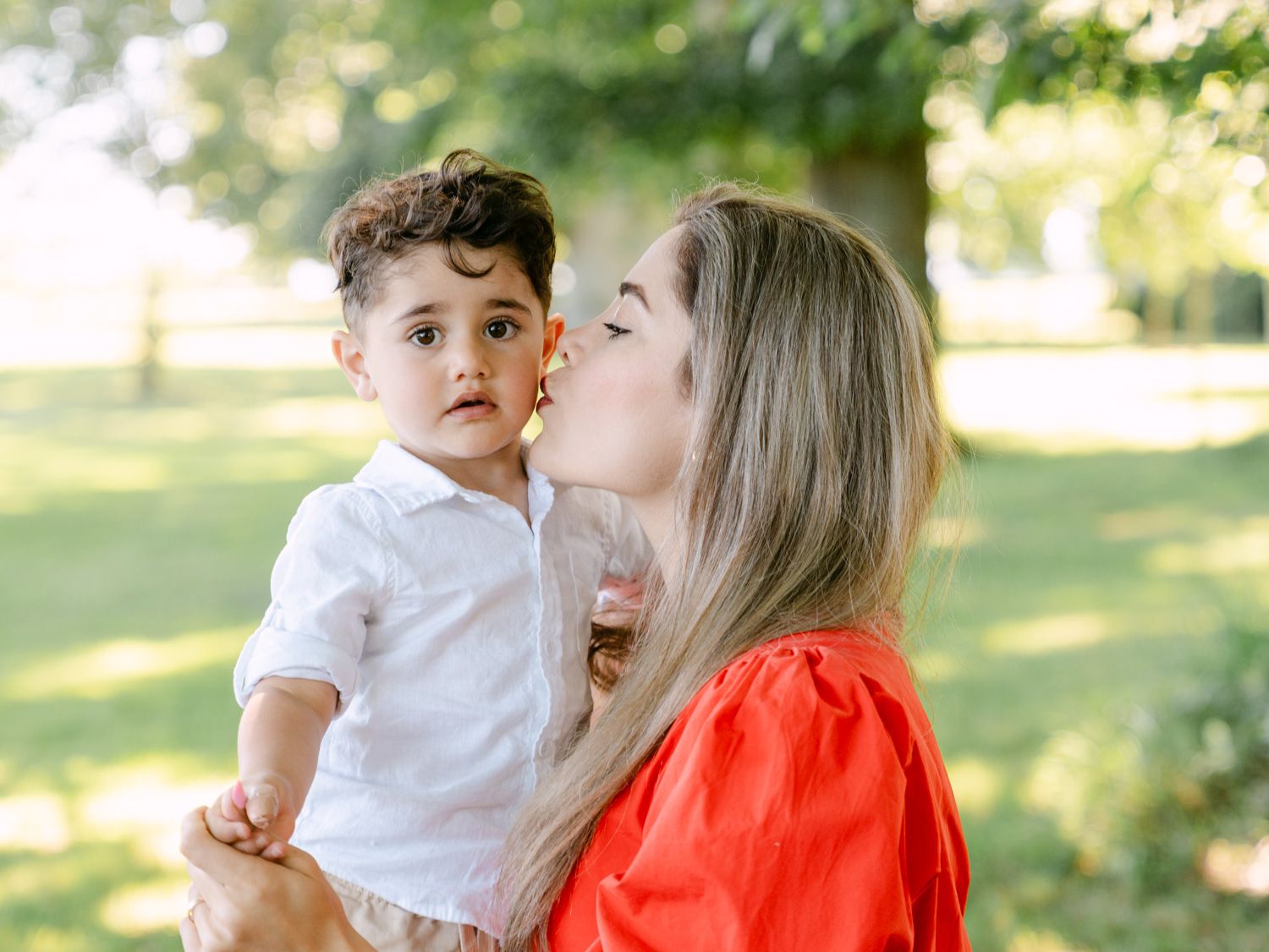 A mother in a red dress kisses her toddler boy in a white shirt outdoors in a lush green park.