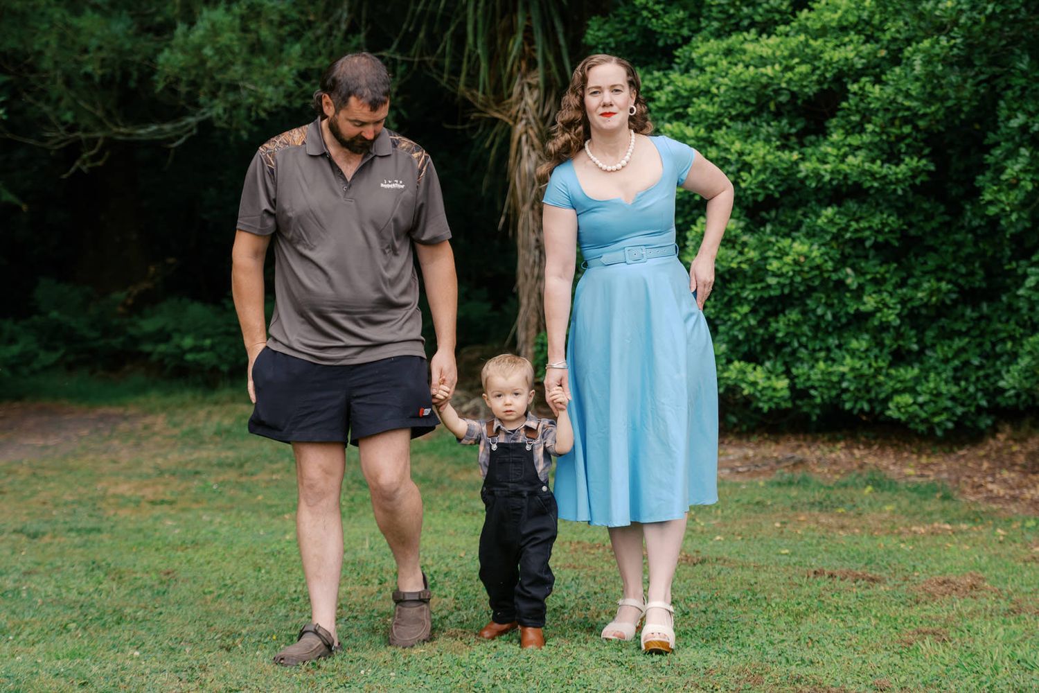 Family of three posing outdoors, parents holding toddler's hands in black overalls, green trees in background.