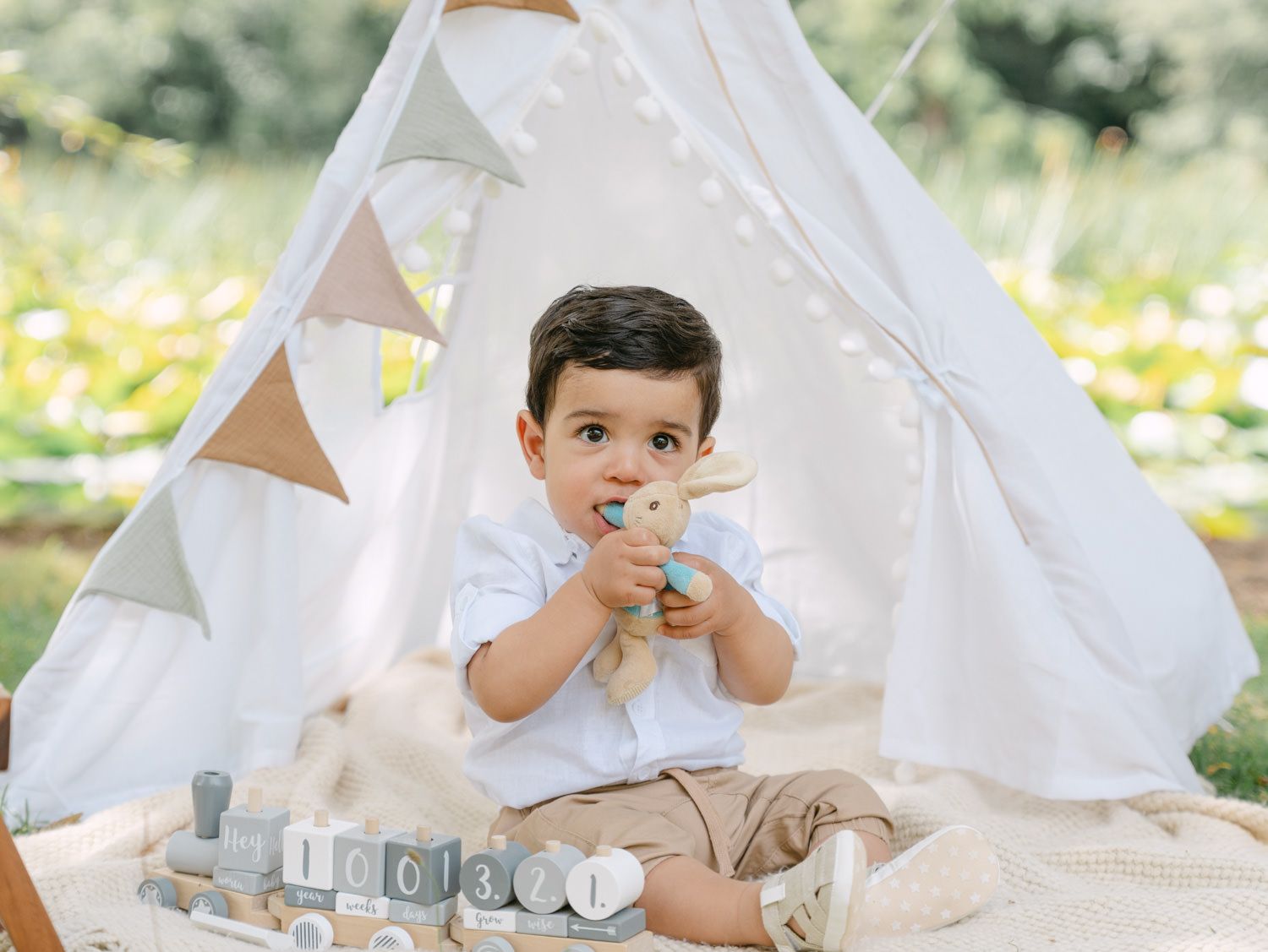 Toddler boy sitting on blanket chewing toy in front of white teepee tent outdoors with soft natural lighting.