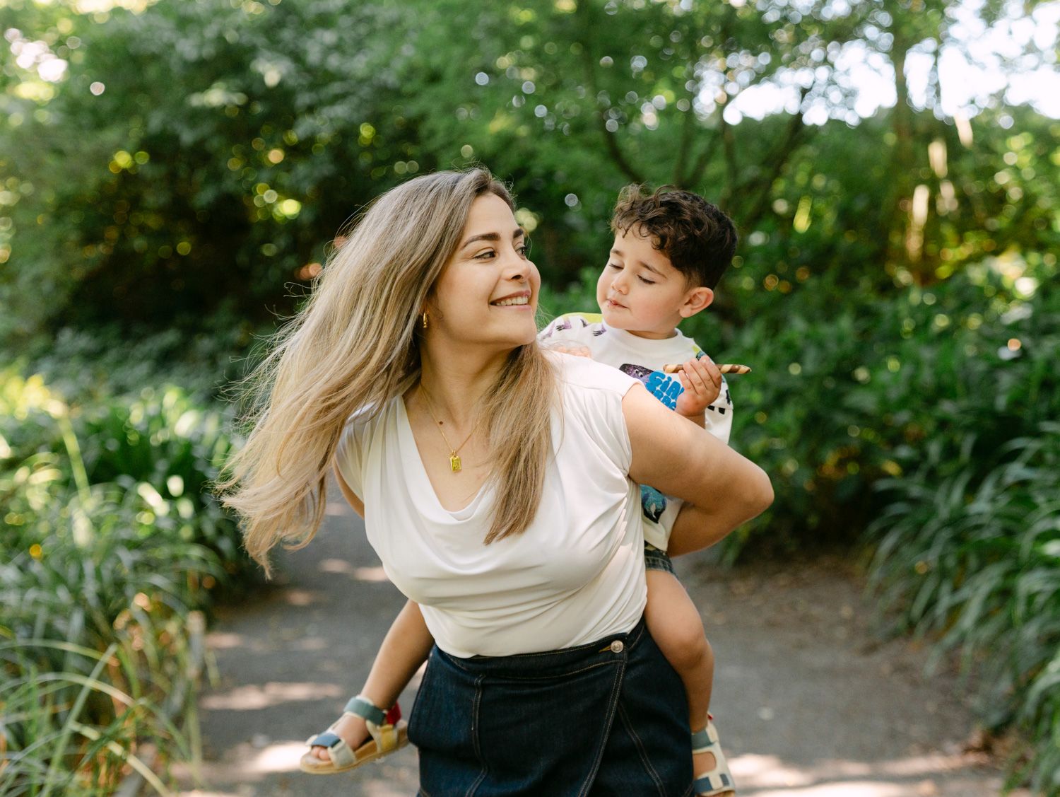 A woman with blonde hair carries a young boy on her back along a lush green garden path.