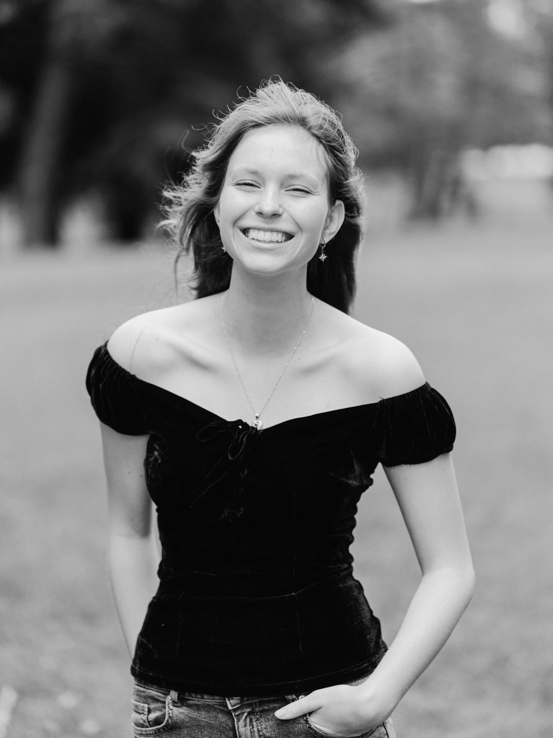 Young woman smiling outdoors in black off-shoulder top, black and white portrait photo.