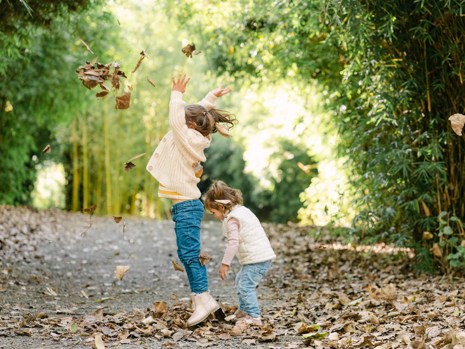 Two young girls joyfully tossing autumn leaves on a tree-lined path in a lush green forest setting.