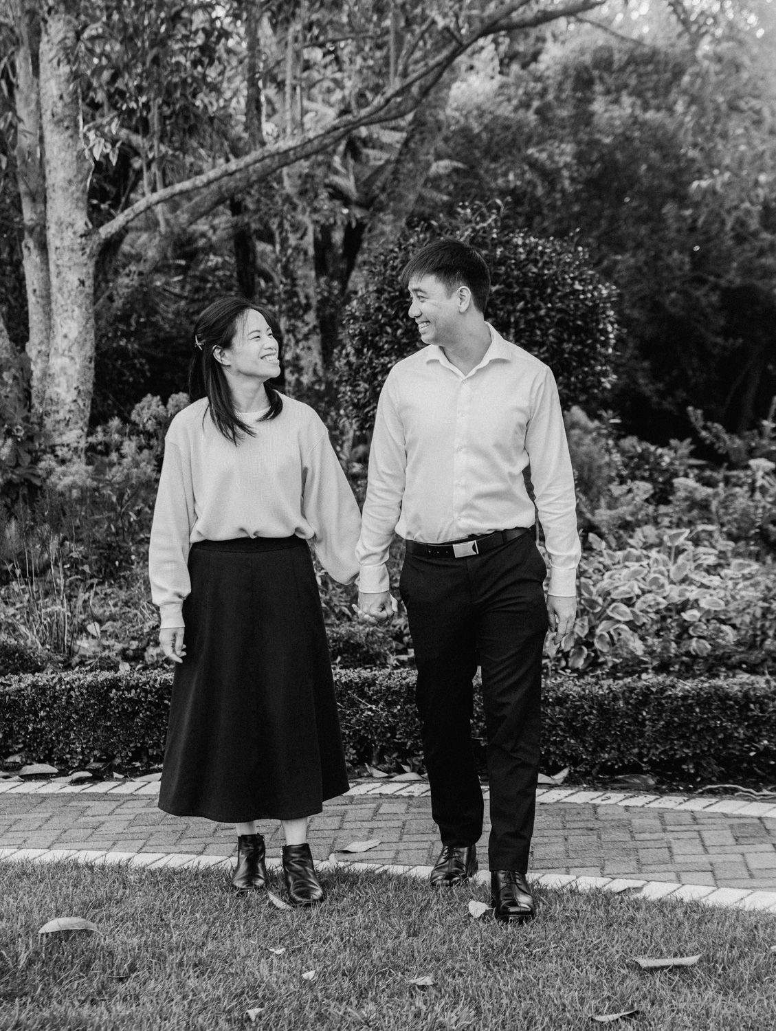 Black and white photo of a couple holding hands and smiling at each other while walking in a lush garden.