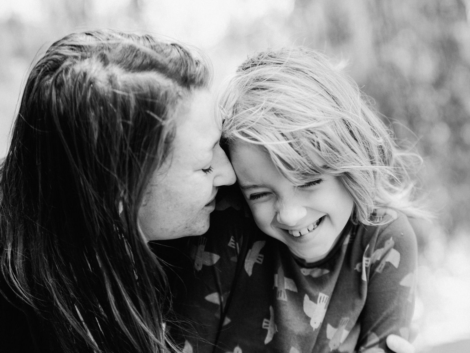 Black and white photo of a mother whispering to her laughing young daughter outdoors.