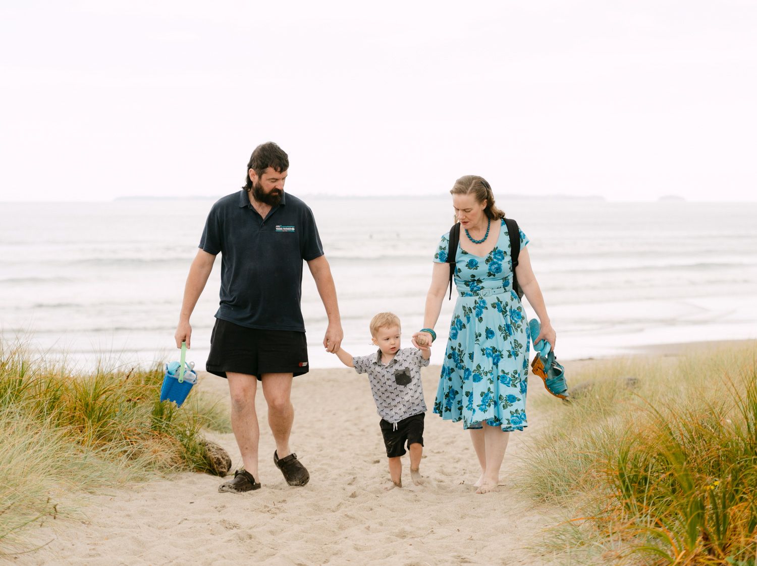 Family of three walking hand-in-hand on a sandy beach path, parents and toddler enjoying a seaside stroll.