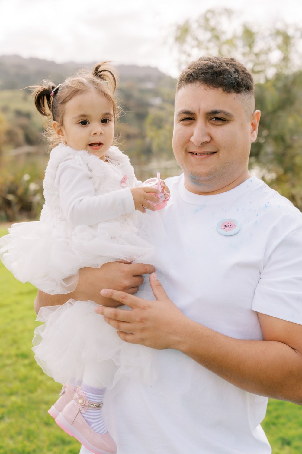 A man in a white t-shirt holds a toddler girl in a white tutu dress outdoors on a green lawn.