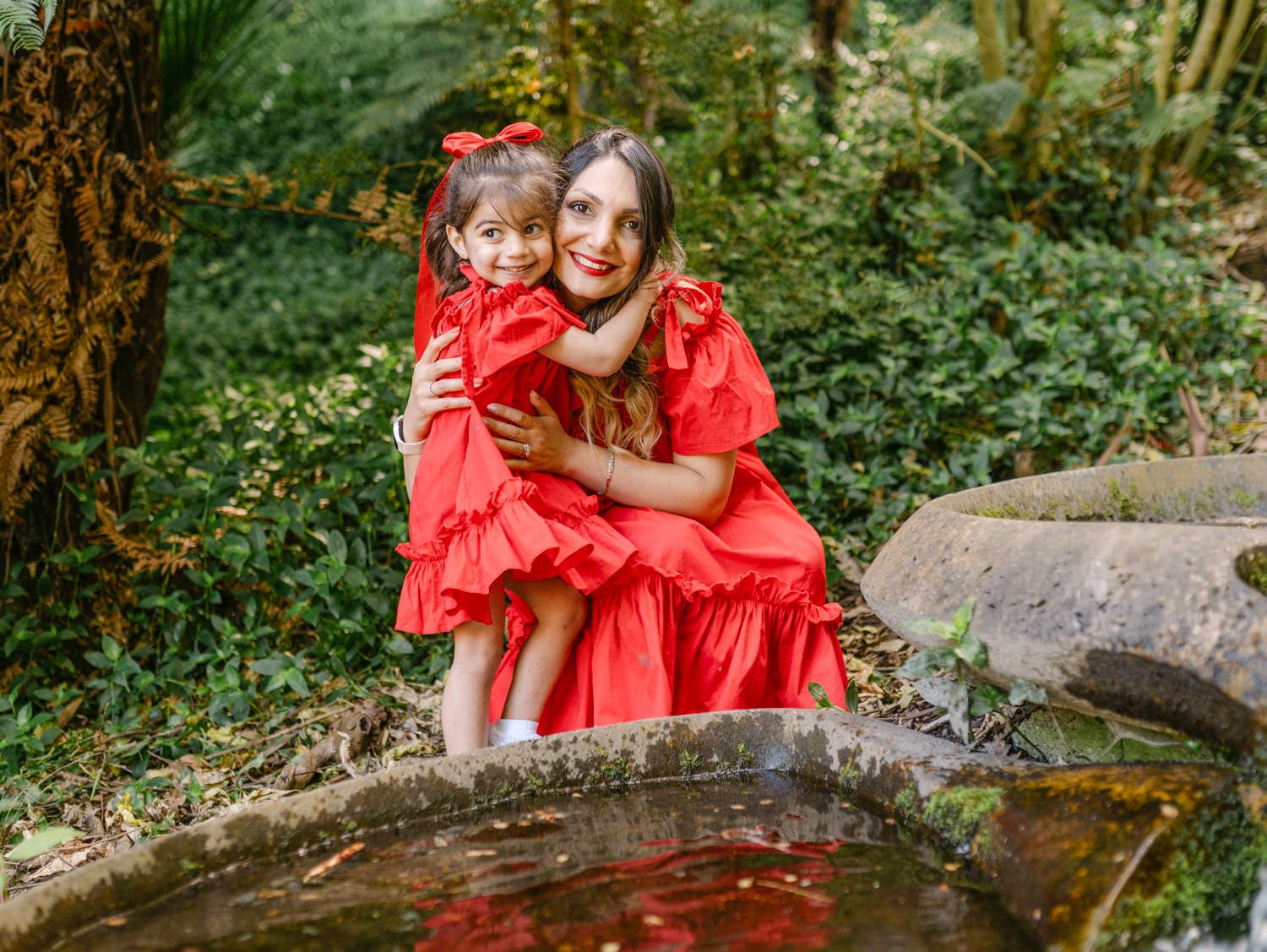 Mother and daughter in matching red dresses hugging outdoors near a pond surrounded by lush greenery.