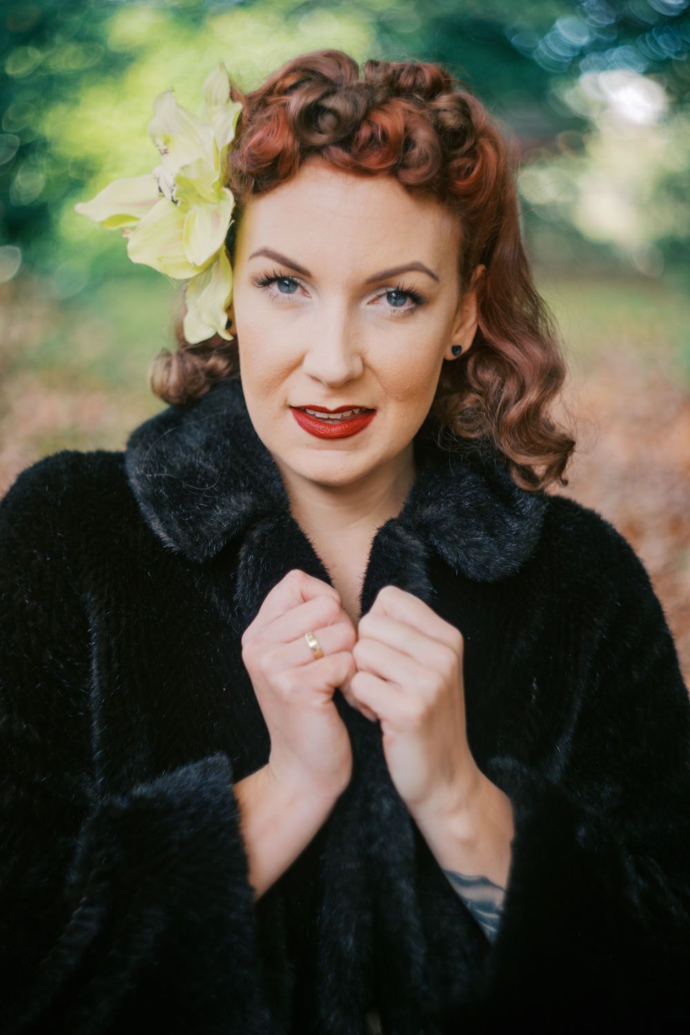Vintage pin-up woman with red curls, yellow flower hair accessory, red lips, and black fur coat posing outdoors.