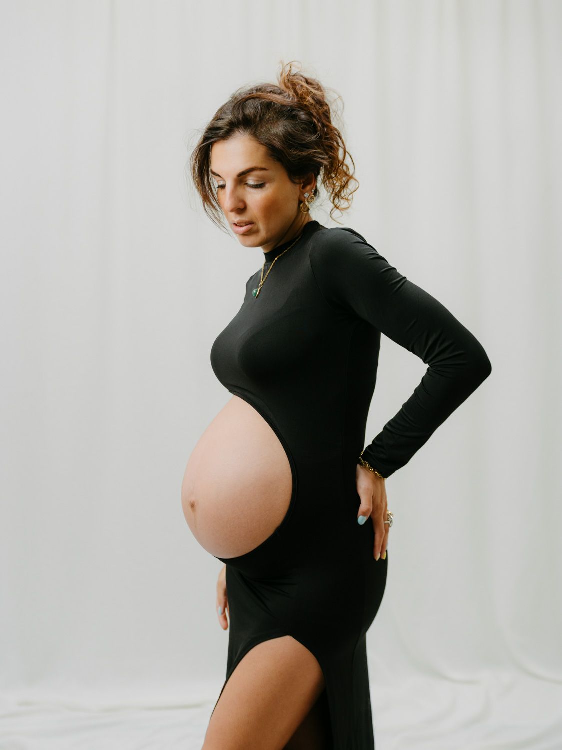 Pregnant woman in black long-sleeve cutout dress posing sideways, showcasing baby bump in a bright studio setting.