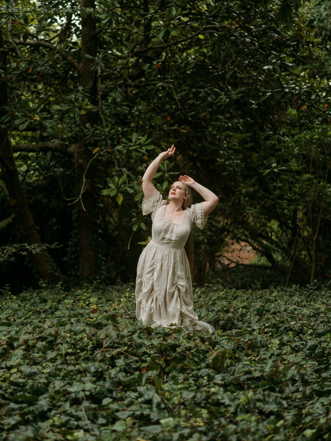 A woman in a vintage white lace dress stands gracefully among lush green ivy in a woodland garden.