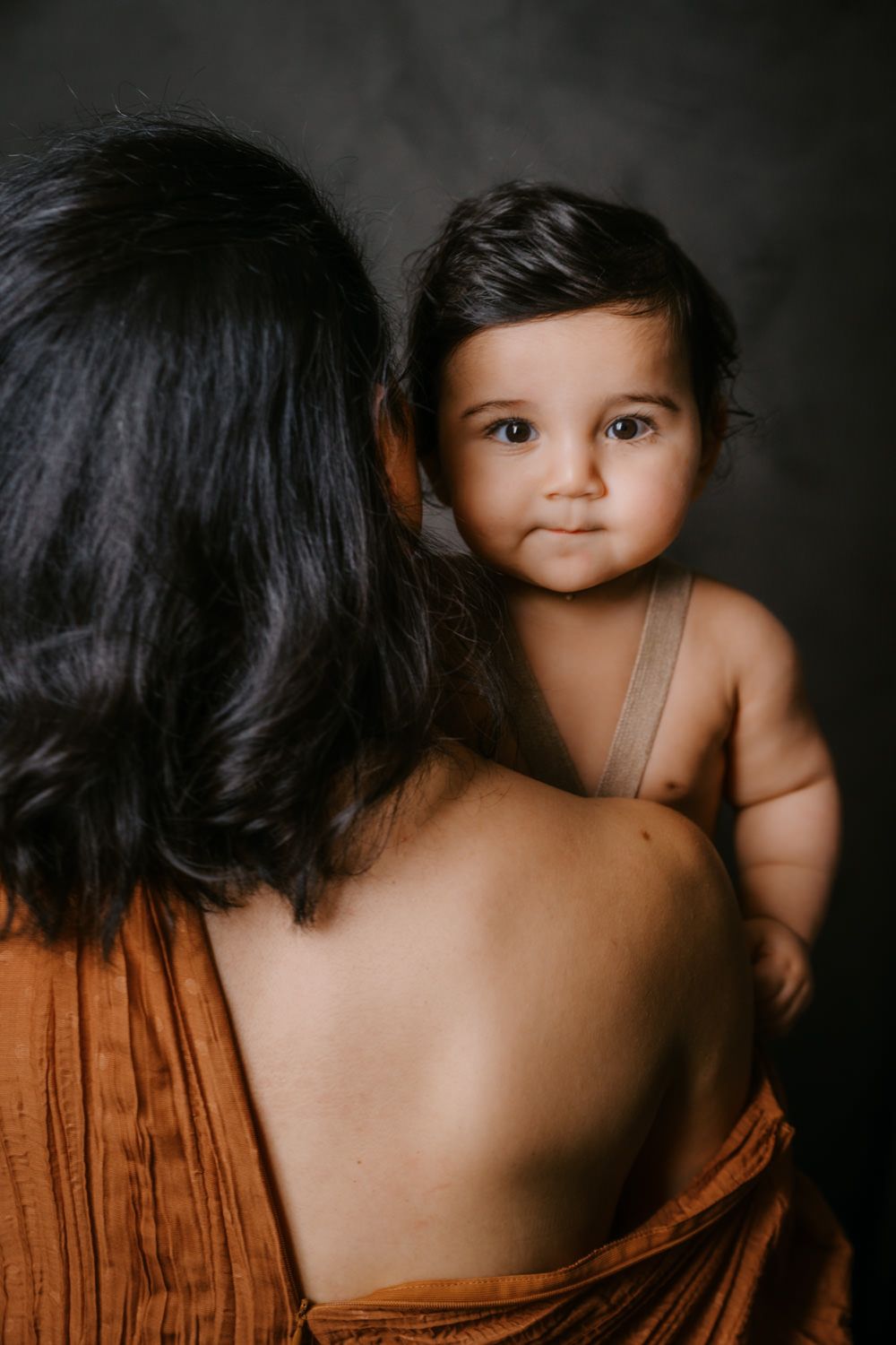 A mother in a rust-colored dress holds her dark-haired baby, who gazes at the camera against a dark background.