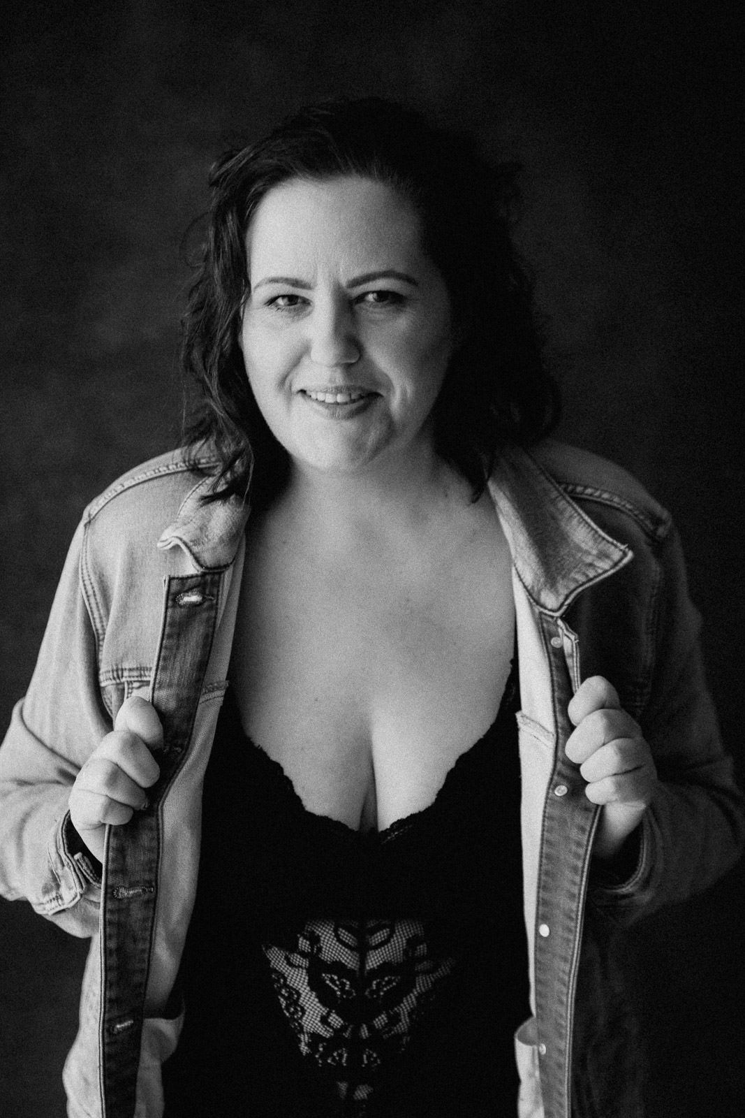 Black and white portrait of a smiling woman in denim jacket and lace bodysuit posing confidently against dark background.