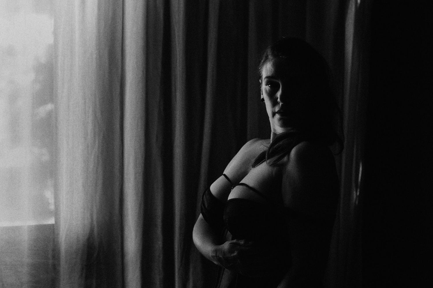 Black and white maternity photo of pregnant woman in lingerie standing by curtained window in soft light.