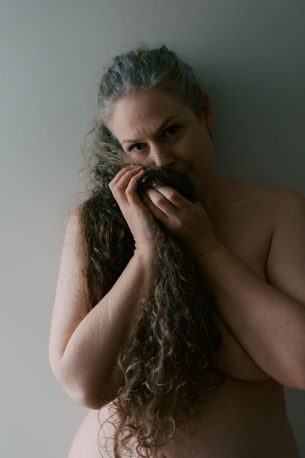 A shirtless person with long curly gray hair covering their face with their hands against a gray wall.