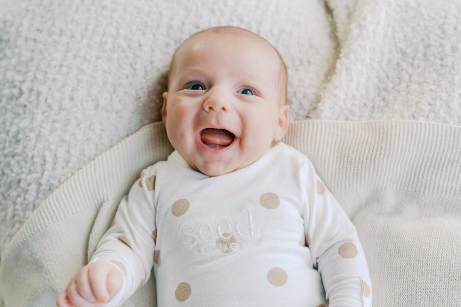 Happy smiling baby lying on a soft white blanket wearing a polka dot onesie.