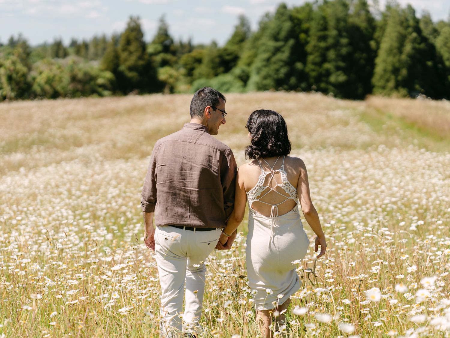 Couple walking hand in hand through a blooming daisy field with lush green trees in the background.