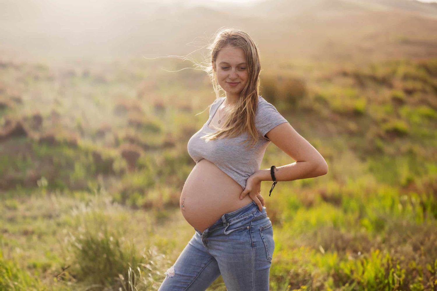 Smiling pregnant woman in gray top and jeans posing outdoors in sunlit green hills, showing baby bump.
