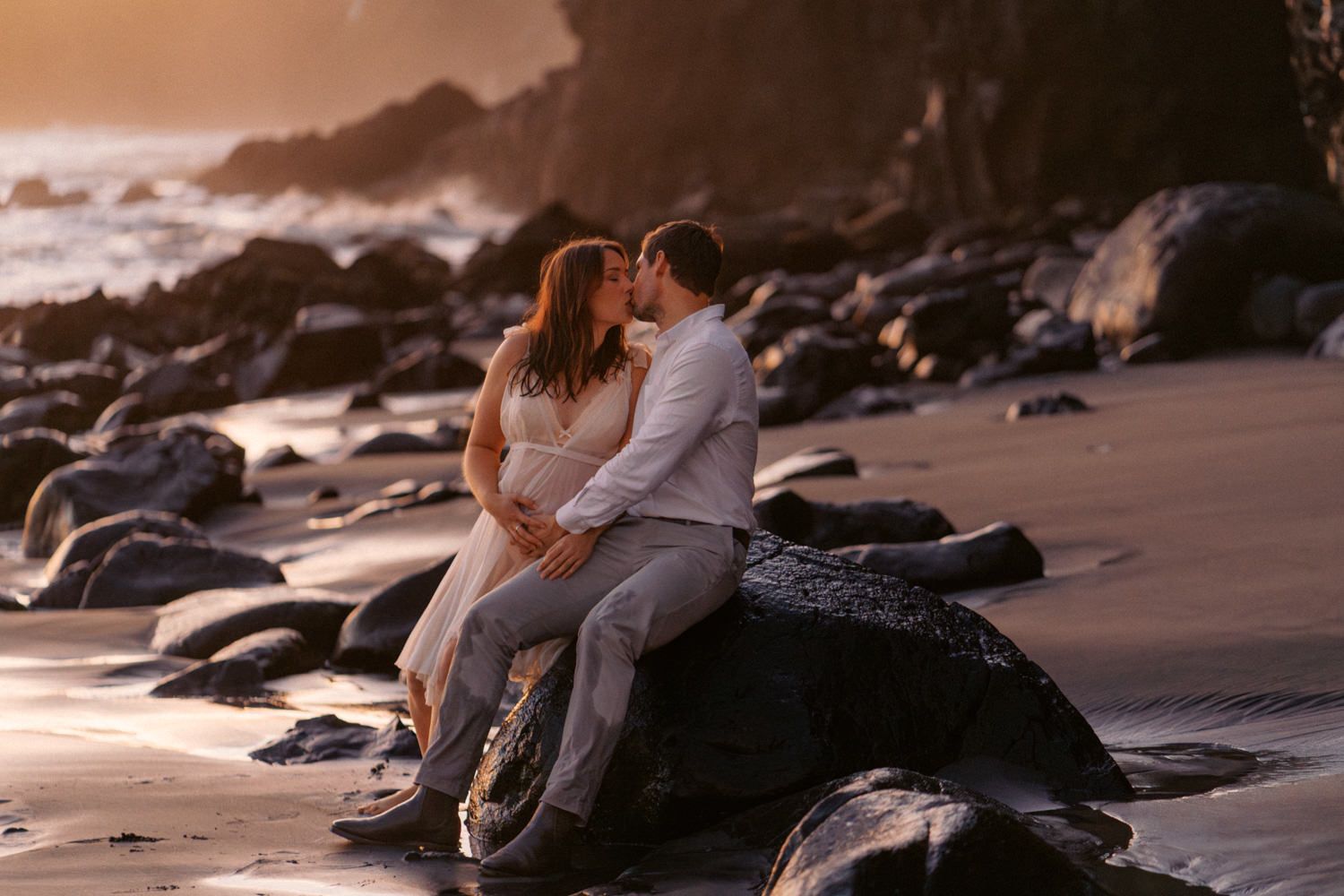 Romantic couple sharing a kiss while sitting on rocks at a sandy beach during a warm golden sunset.
