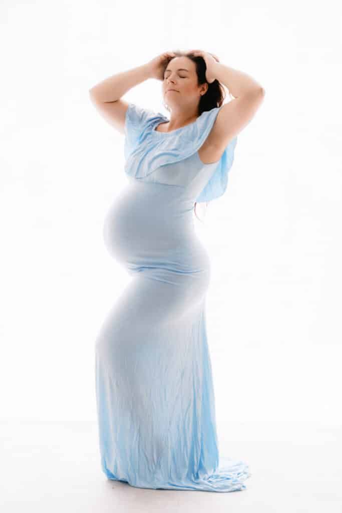 Pregnant woman in flowing light blue gown poses gracefully with hands in hair against white background.
