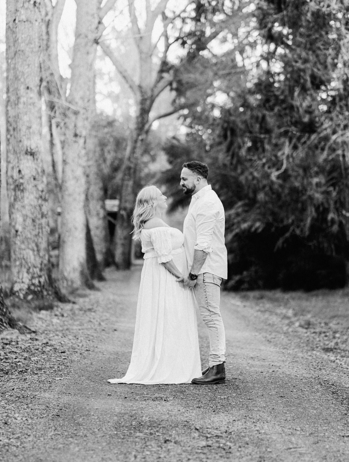 Black and white photo of a couple holding hands on a tree-lined path, woman in flowing white gown, man in casual attire.