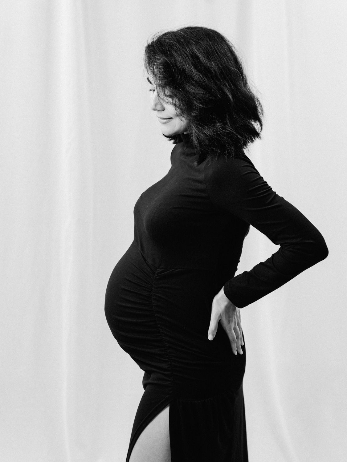 Black and white maternity photo of pregnant woman in black turtleneck dress posing sideways against white backdrop.