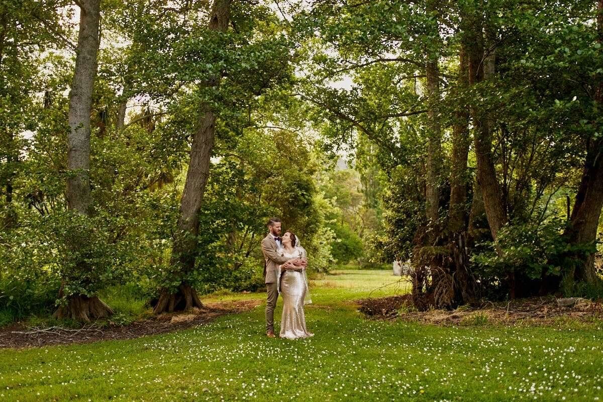 Bride and groom embracing in a lush green woodland garden, surrounded by tall trees and wildflowers.