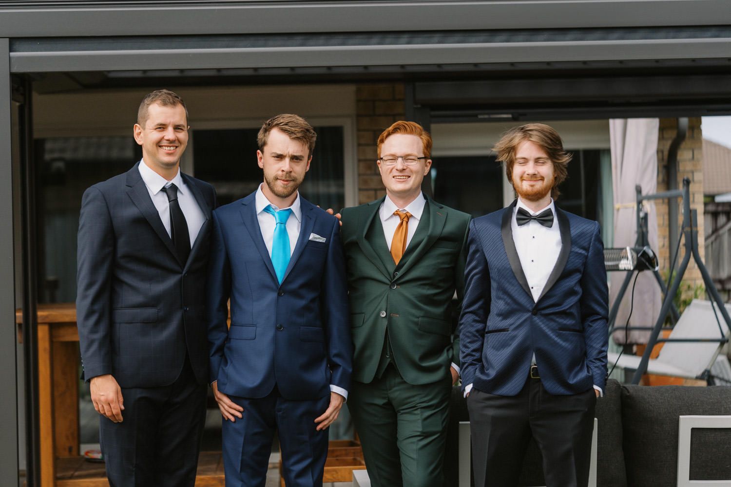 Four well-dressed men in suits smiling together outdoors at a formal event or wedding celebration.