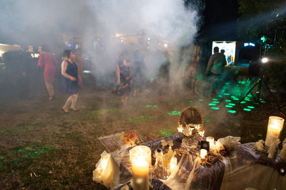 Outdoor night party with candles, smoke effects, and green lighting on grass with guests dancing in background.