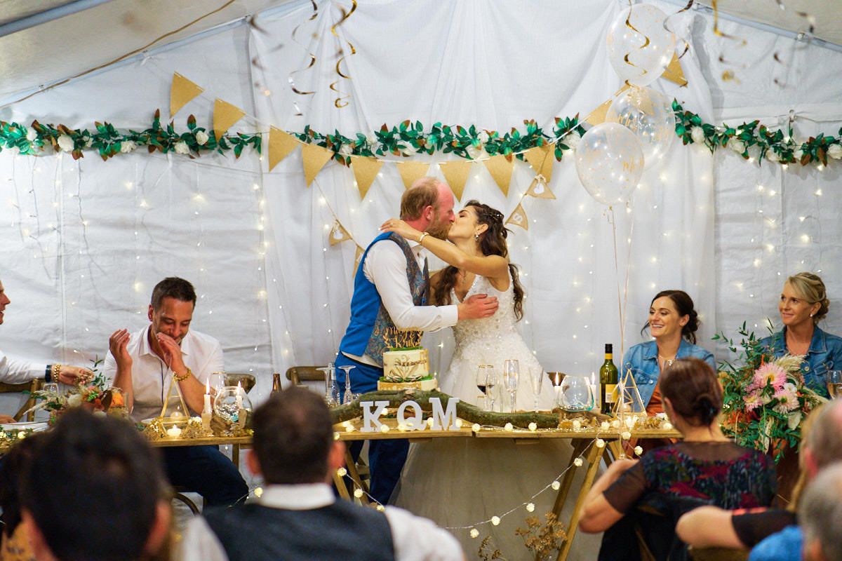 Bride and groom share a kiss at wedding reception table in a marquee with gold and green decorations.