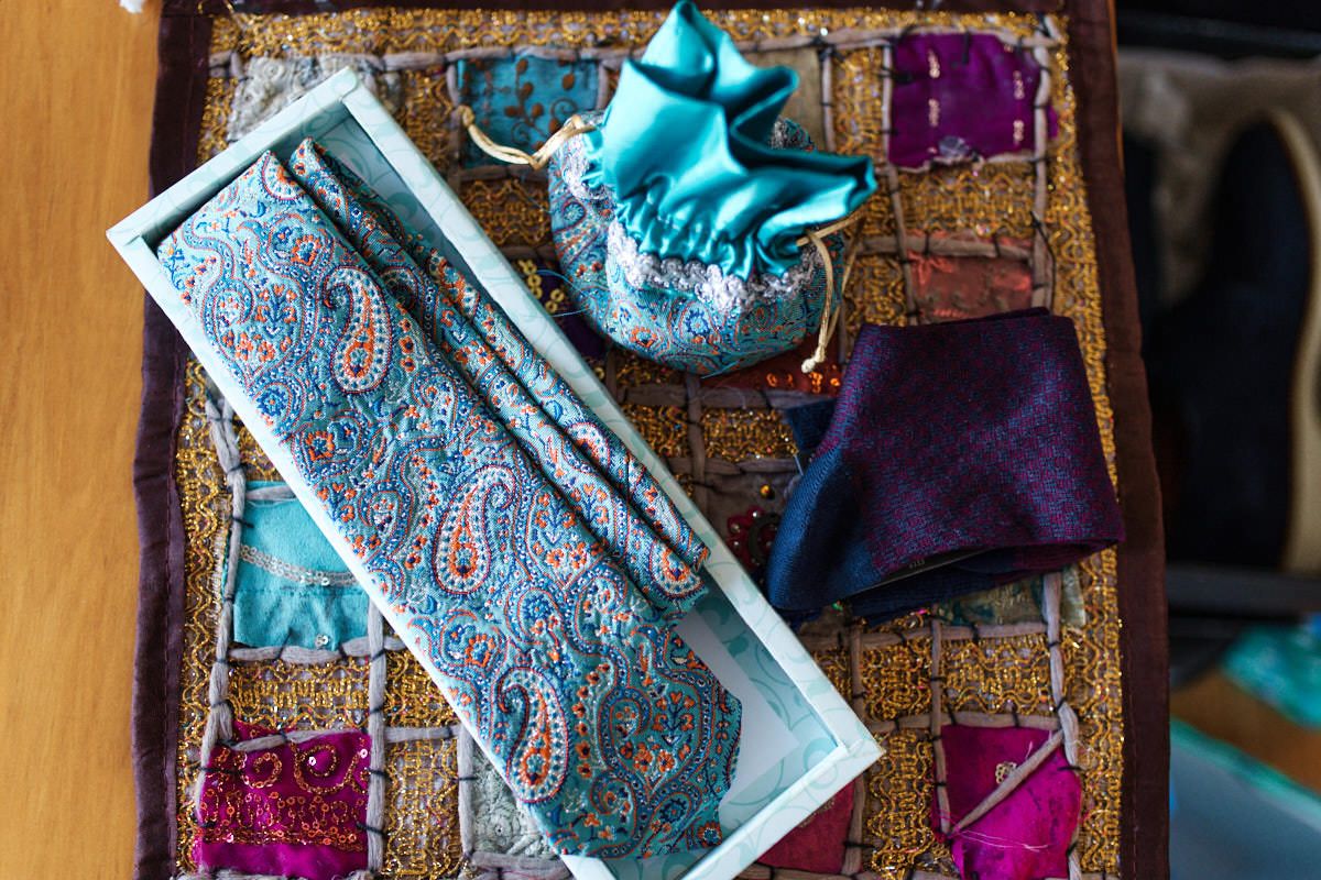 Teal paisley tie in white box with purple pocket square and satin pouch on ornate patchwork fabric.
