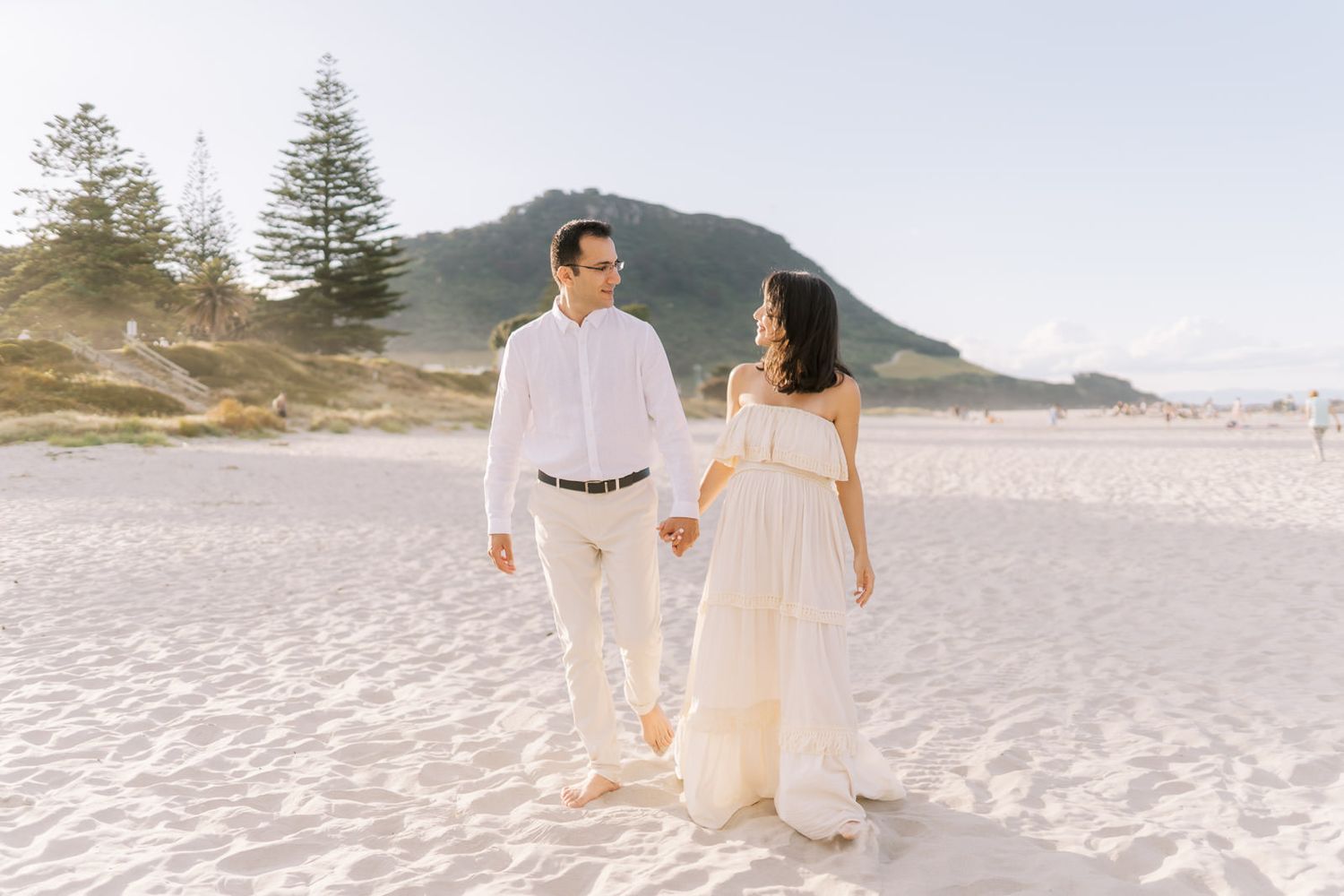 Couple walking hand in hand on a sandy beach in white outfits, with a scenic mountain backdrop.