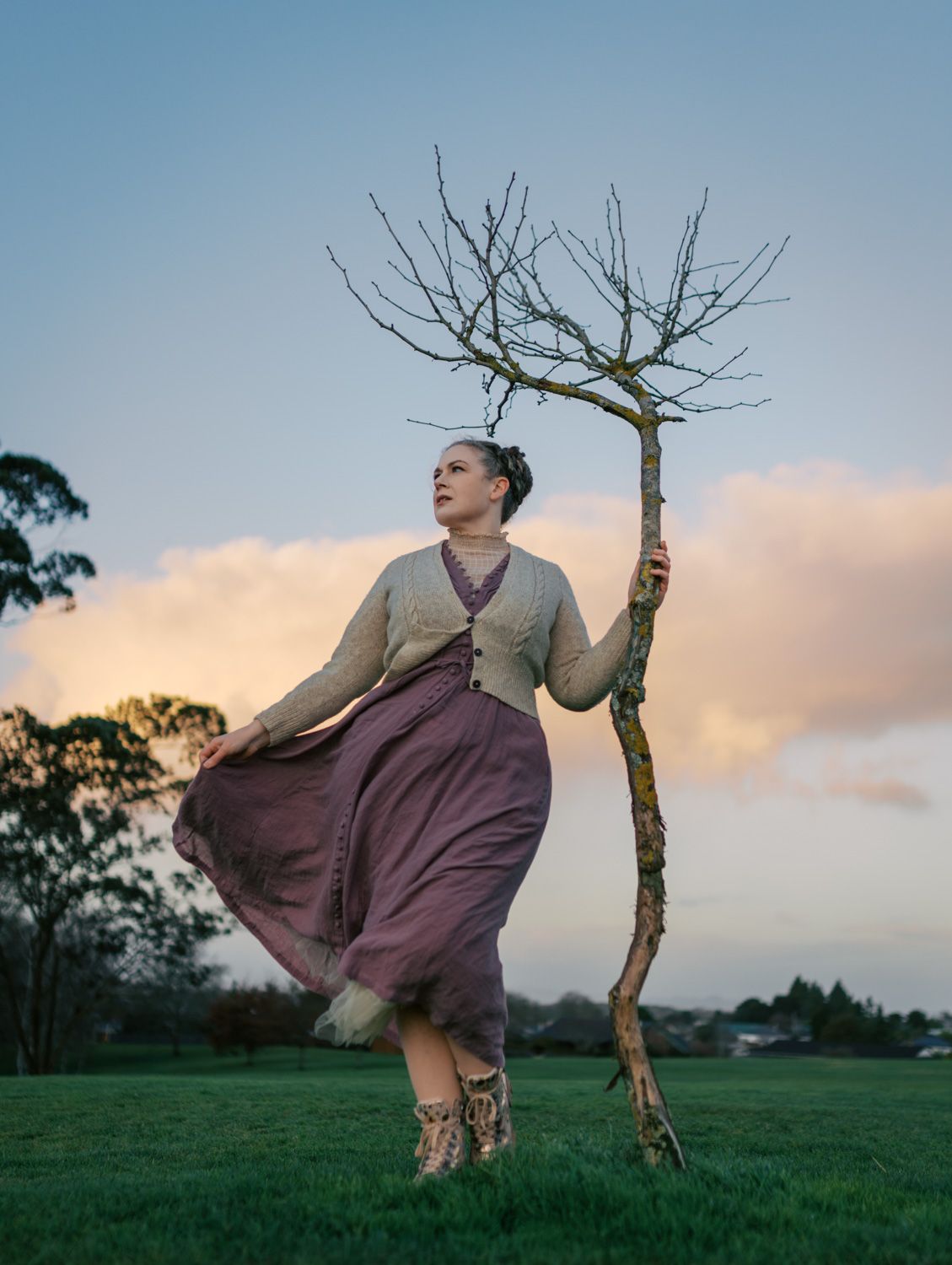 Woman in mauve skirt holding bare tree branch in green field at dusk with dramatic cloudy sky.