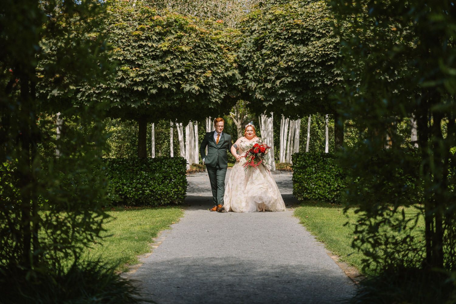 Bride and groom walking hand in hand through an elegant tree-lined garden pathway on their wedding day.