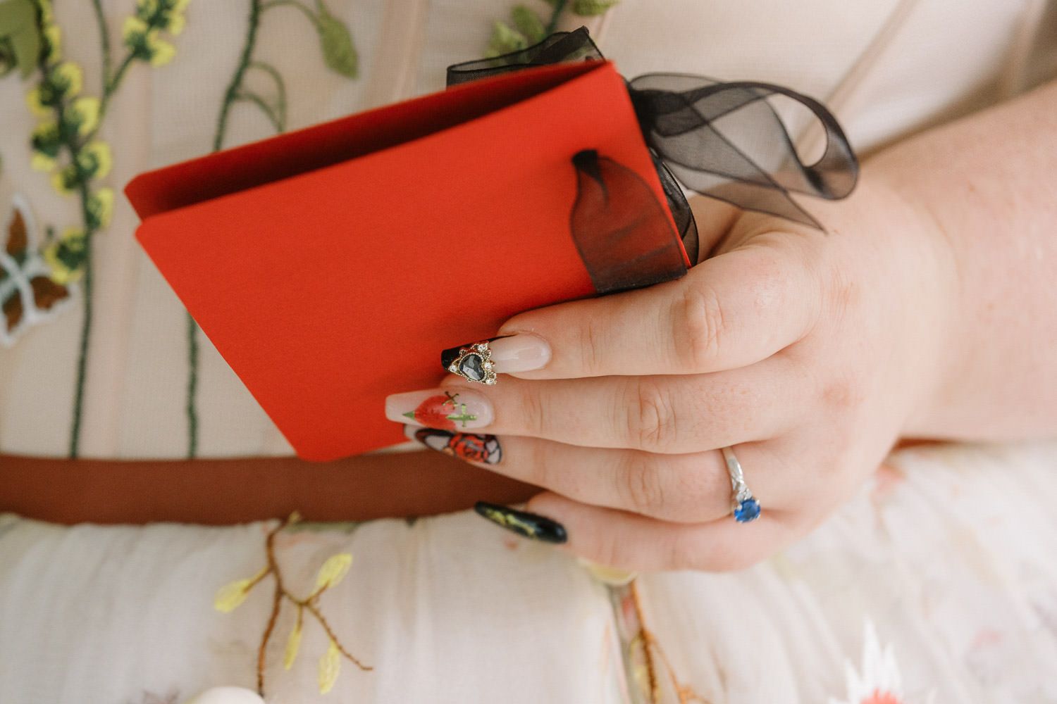 Close-up of a bride holding a red clutch purse with decorated nails and a blue gemstone ring.