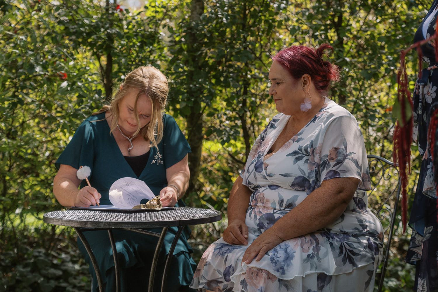 Two women signing wedding documents at outdoor table surrounded by lush greenery.