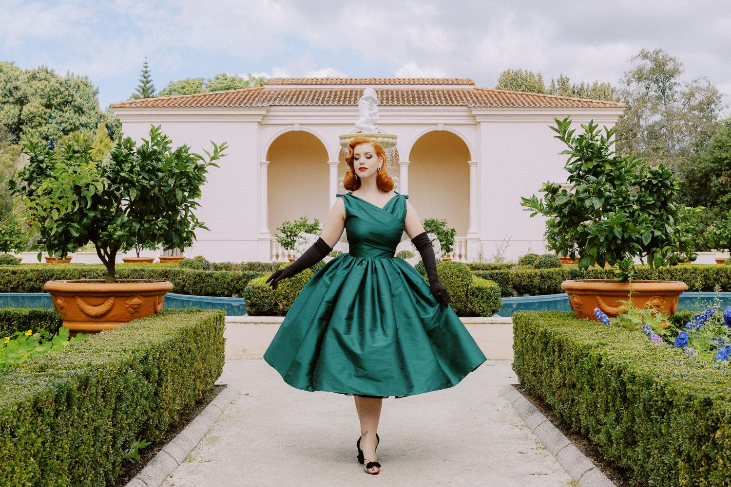 Woman in green 1950s dress with black gloves poses in elegant garden with hedges and white building.