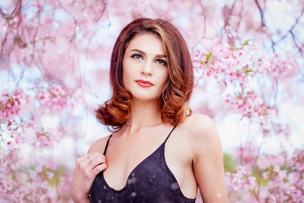 Young woman with auburn hair poses in black dress among blooming pink cherry blossom trees in spring.